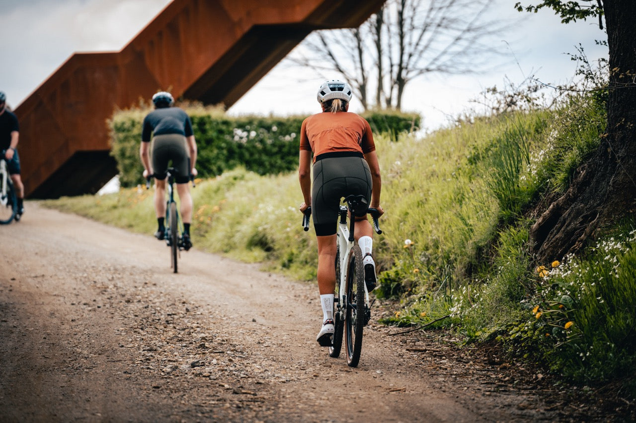 Close-up rear view of a cyclist wearing Bioracer white performance cycling socks while riding uphill on a gravel path, highlighting fit, compression and race-ready comfort.
