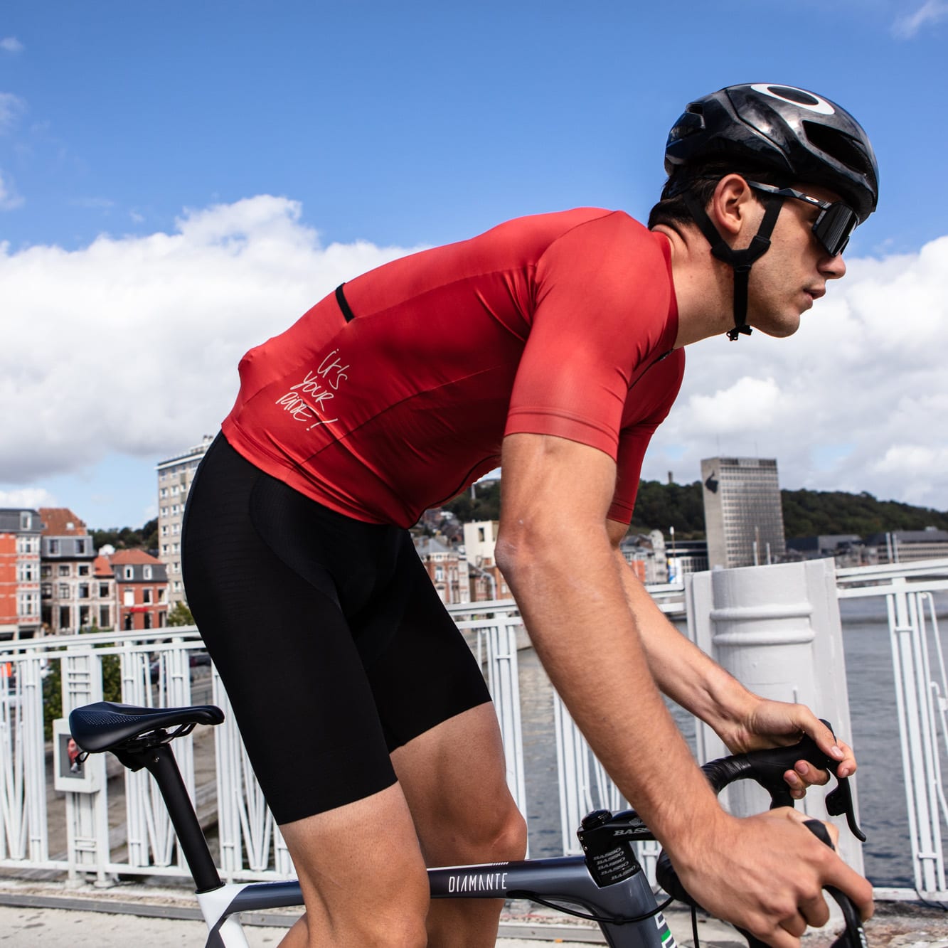 Male cyclist wearing the red Bioracer Metalix Jersey with 'It's Your Ride' text, riding a road bike across a city bridge on a sunny day