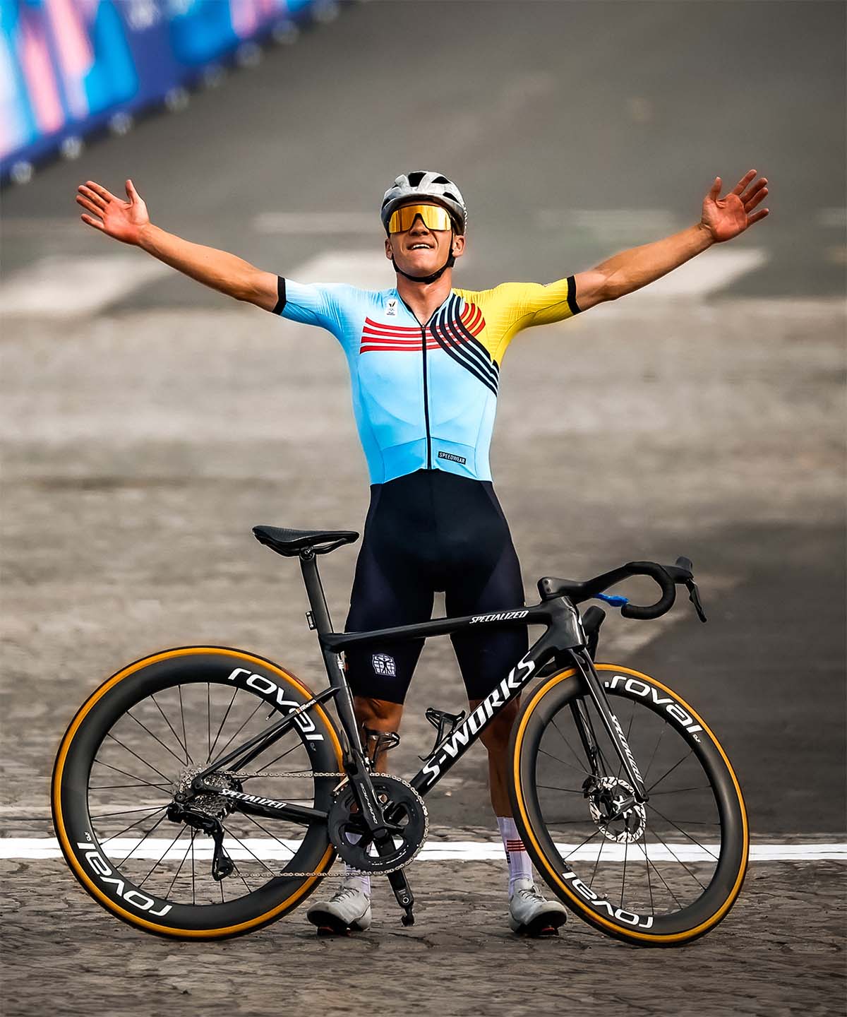 Remco Evenepoel celebrates with arms wide open after a solo victory, standing behind his black S-Works bike, wearing the Belgian national time trial champion kit.