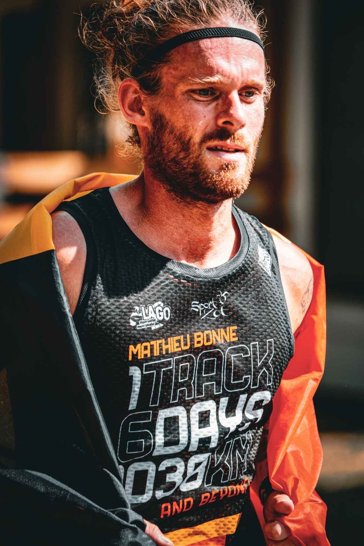 Close-up of ultra runner Matthieu Bonne wearing a Belgian flag over his shoulders after completing his 6-day, 1038 km track challenge, dressed in a black mesh race tank with bold “TRACK 6 DAYS 1038 KM AND BEYOND” lettering.