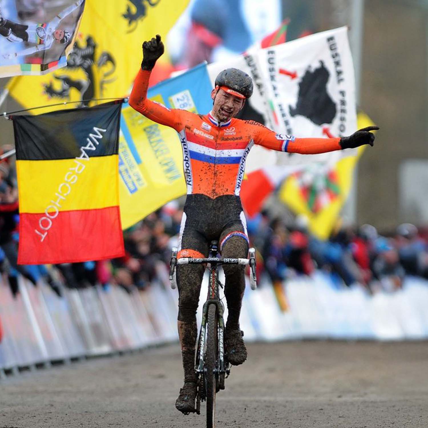 Mathieu van der Poel crossing the cyclocross finish line covered in mud, celebrating victory in the Dutch national kit, with Belgian and Dutch flags waving in the background.