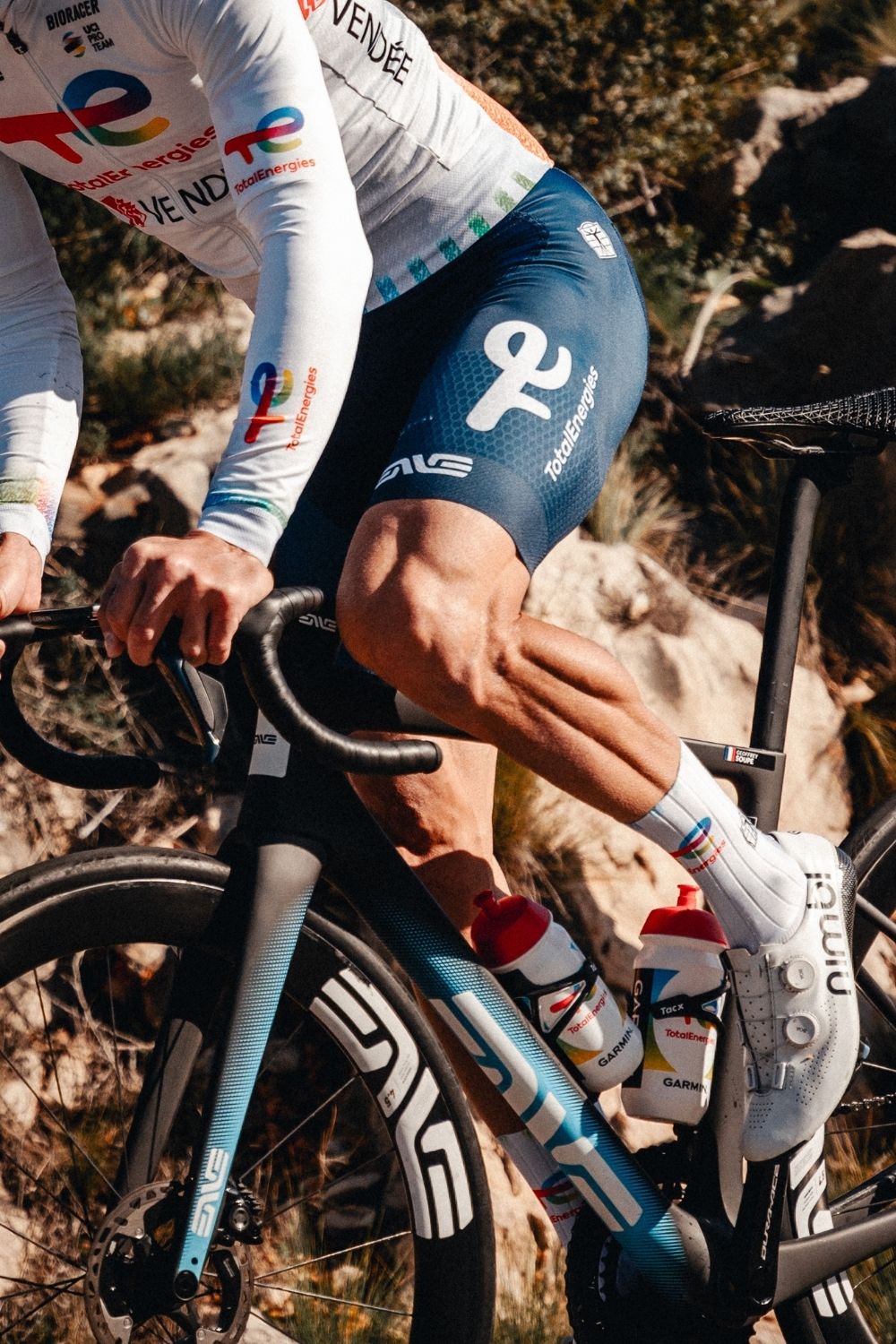 Close-up of a Team TotalEnergies pro cyclist riding an ENVE road bike, showcasing muscular legs, branded cycling kit by Bioracer, and dual water bottles with TotalEnergies and Garmin logos.