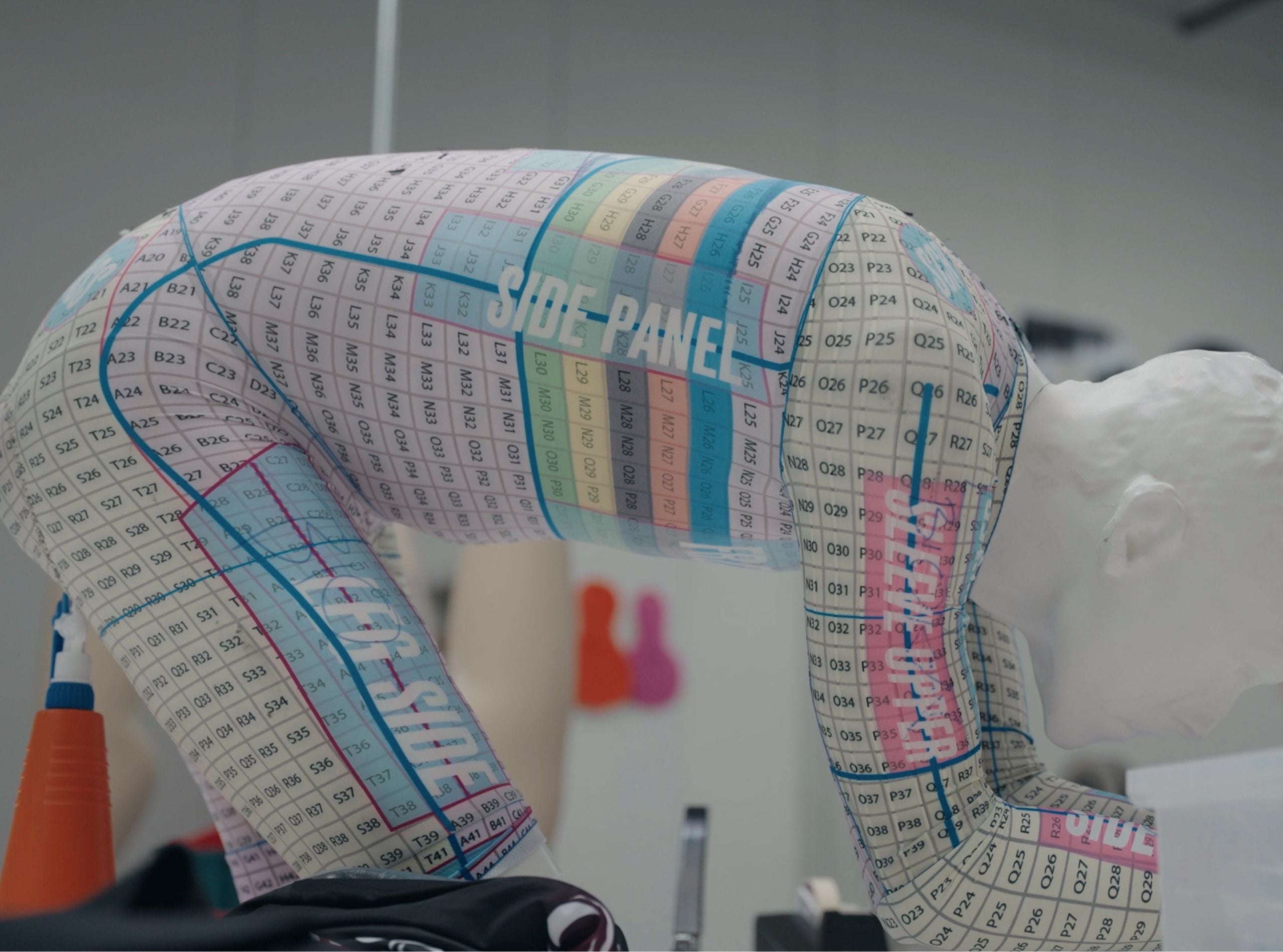 White mannequin in aerodynamic time trial position, wearing a Bioracer sizing suit marked with grid codes and color zones for individual pattern development and panel fitting.