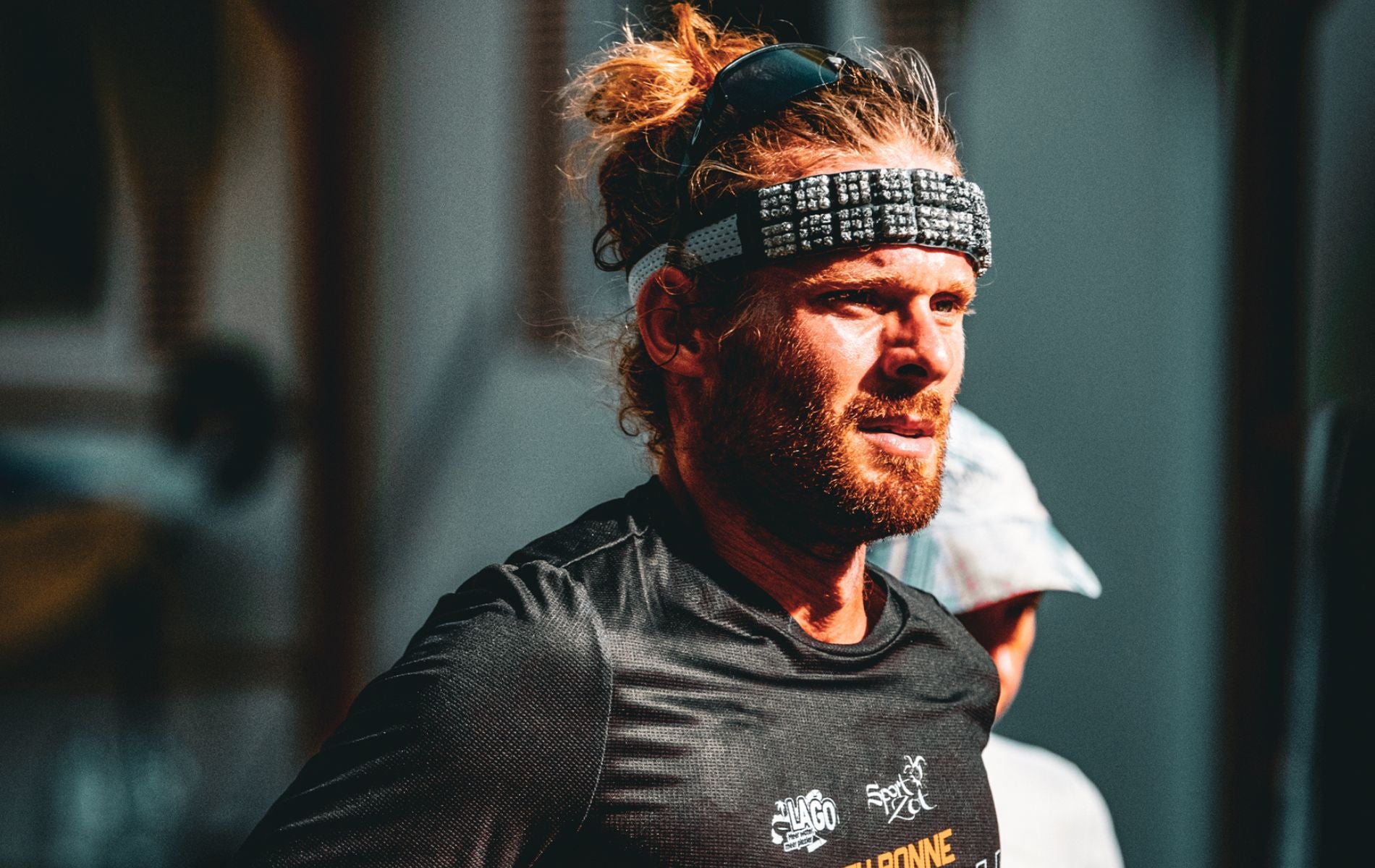 Determined ultra runner Matthieu Bonne mid-race in the sun, wearing a black performance shirt and patterned headband, with hair tied back.