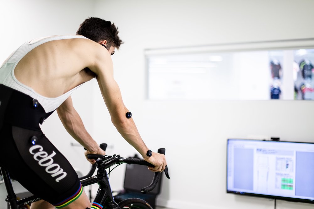 Wout van Aert undergoing a professional bike fit session, wearing motion capture sensors while riding indoors on a race bike, with a performance analysis screen visible in the background.